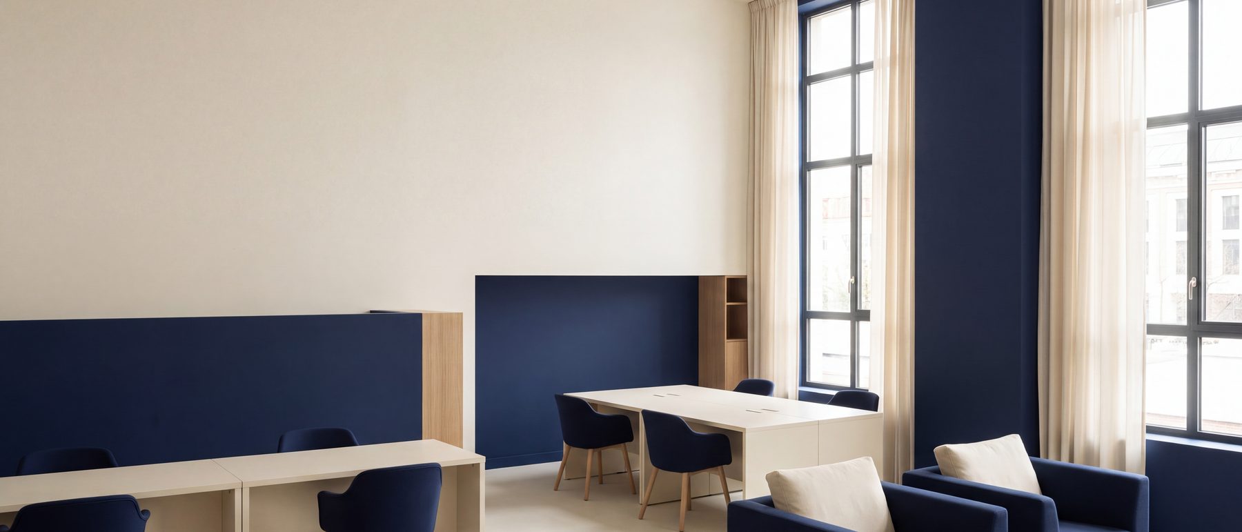 Bureau aux murs crème et panneaux marine, postes de travail et aire de détente sous de hautes fenêtres